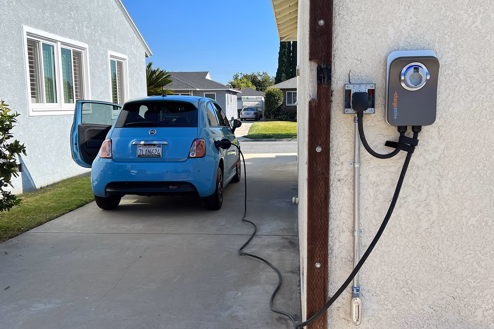 Blue Fiat 500e electric car parked near a wall-mounted EV charging station with a charging cable connected.