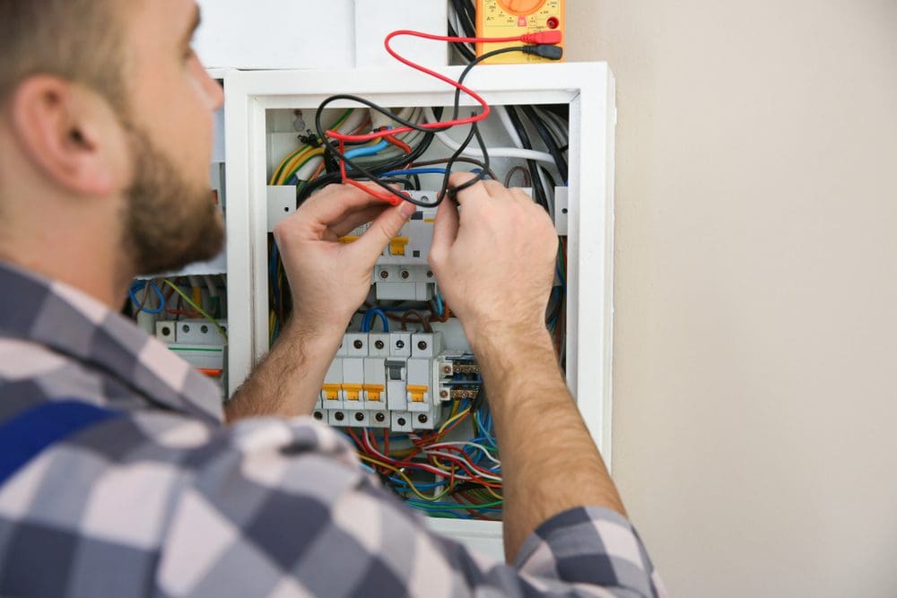 Electrician working on an electrical panel, connecting wires and checking connections with tools in a residential setting.