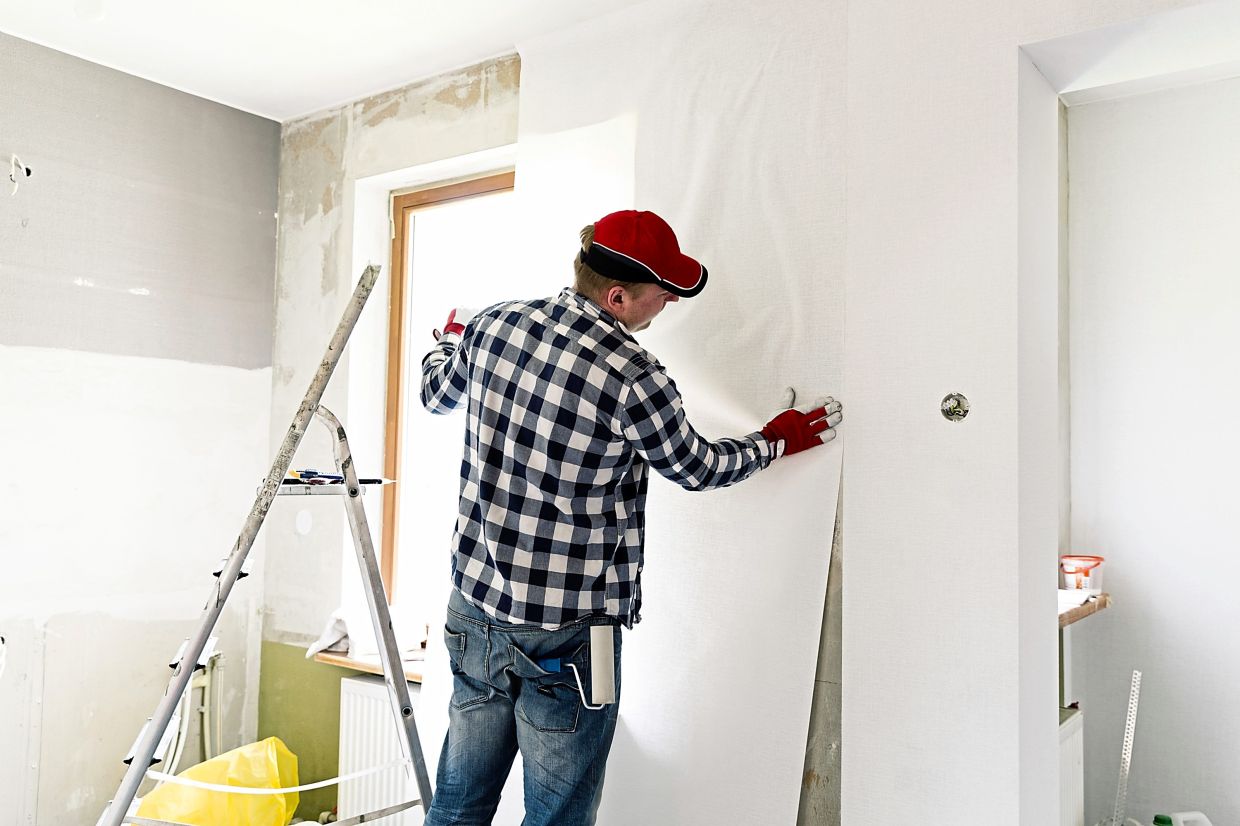 A worker applying wallpaper in a room, wearing gloves and a cap, while standing on a ladder. Tools and materials are visible nearby.