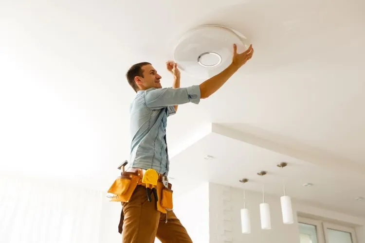 Man installing a ceiling light fixture while standing on a ladder, wearing a tool belt with various tools.