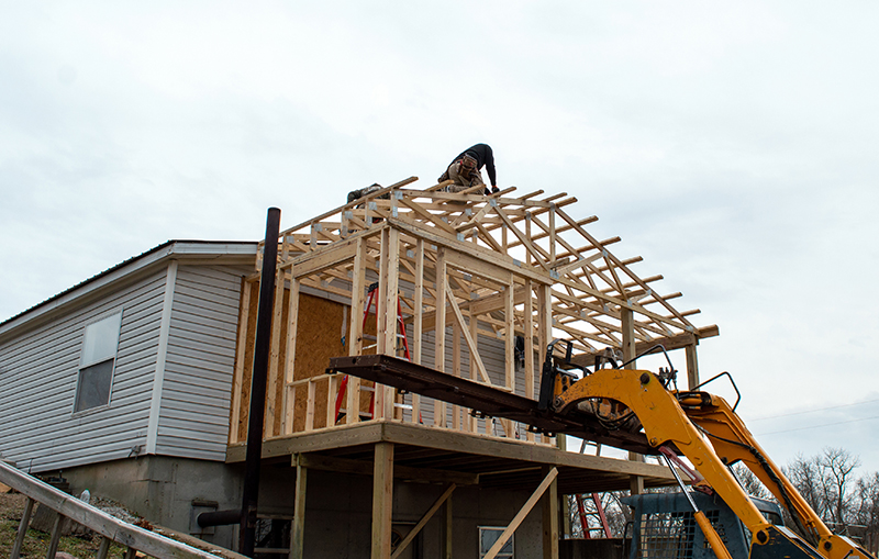 Construction of a wooden frame for a home addition, with a worker on the roof and machinery nearby.